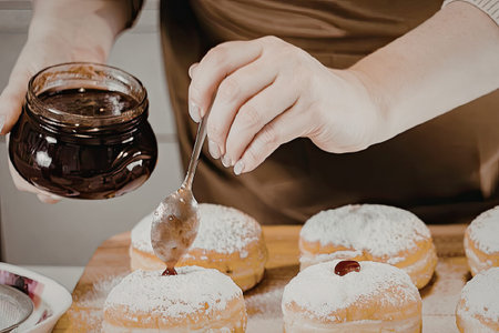 A person in a brown apron spreading jam on donuts dusted with powdered sugar.