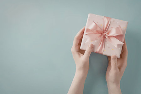 A pair of hands holding a pink gift box with a ribbon tied around it on a light blue background.
