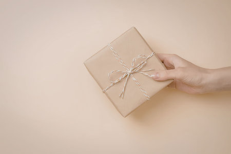 A hand holding a square gift box wrapped in brown paper and tied with twine on a beige background.