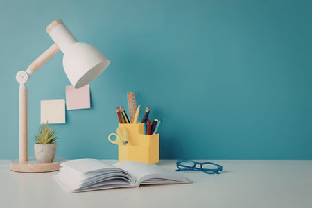 A study desk with a lamp, a plant, a book, glasses, and a container with pens and pencils.