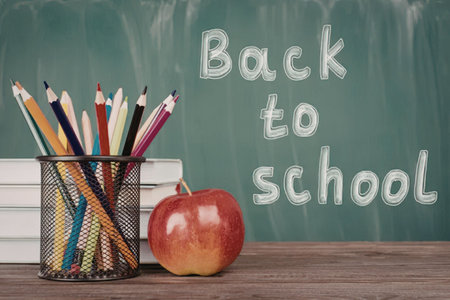 A black mesh pencil holder filled with colorful pencils and pens, a red apple, and a stack of books...