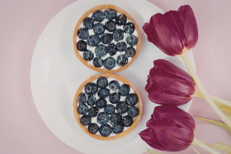 Two blueberry tarts on a white plate with pink tulips on the side