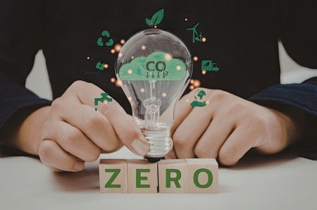 A person holding a lightbulb with a green cloud and CO2 inside, surrounded by green leaves and blocks with the word 'ZERO' on a table.