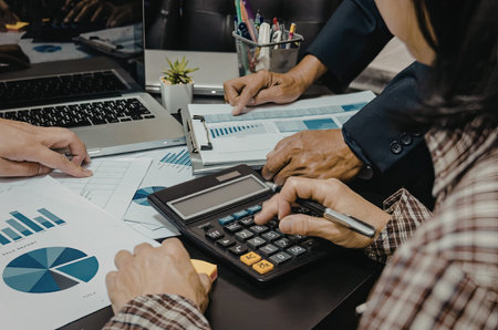 Two people working together at a desk with a calculator, laptop, and papers with charts and graphs.