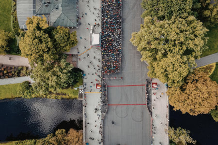 An aerial view of a large group of people participating in a marathon or fun run event. The event is taking place on a road that runs through a park with trees and a body of water on either side. The road is closed to traffic and the participants are running in a group.