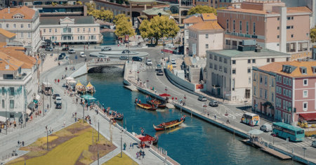 An aerial view of a city canal with boats and buildings along the waterway. The canal is surrounded by a road and sidewalk, with trees and grassy areas nearby. The buildings have orange roofs and are a mix of pastel colors.