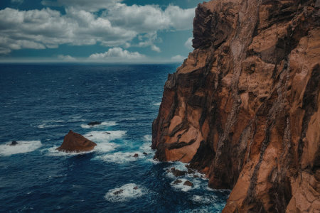 A rugged cliffside stands tall against the crashing ocean waves, with a large rock formation in the sea below, under a blue sky with white clouds.