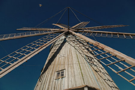 A low angle shot of a windmill with a clear blue sky in the background. The windmill has a weathered wooden exterior and a set of long blades. A small window is visible on the side of the windmill.