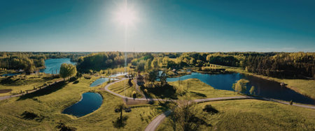 This image captures a breathtaking aerial view of a peaceful park featuring multiple lakes, lush green grass, and walking paths. The serene landscape is set against a clear blue sky with the sun shining brightly.