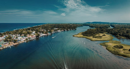 An aerial view of a coastal town with a winding waterway, green islands, and a vibrant blue sea under a partly cloudy sky.
