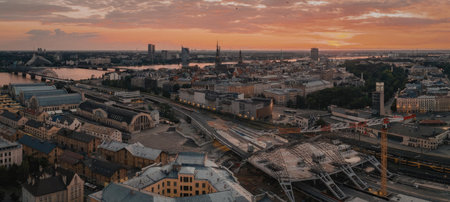 Aerial view of a city with a river running through it during sunset. The city has a mix of old and new buildings, with a prominent road and bridge in the foreground. The sky is painted with hues of orange and pink.