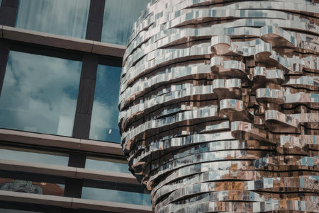 A close-up view of a metallic sculpture with a unique, layered design, situated in front of a modern building with large windows reflecting the sky.