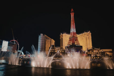 A nighttime view of the Las Vegas Strip featuring a large fountain display in front of a prominent hotel with a tall tower lit up in red and white, surrounded by other buildings and attractions.