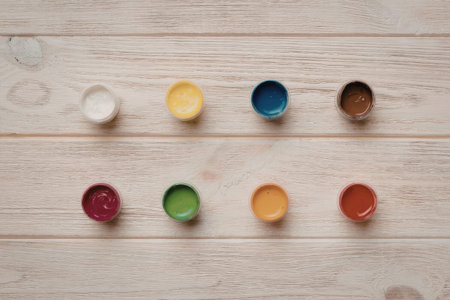 An arrangement of nine paint containers in different colors on a light wooden background, showcasing a variety of hues.