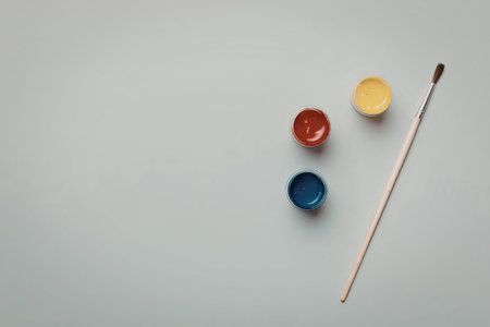 A paintbrush and three small pots of paint in red, blue, and yellow on a gray surface
