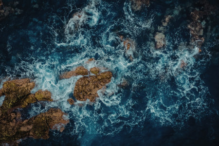 Aerial view of ocean waves crashing against a rocky shoreline with brown rocks and blue water