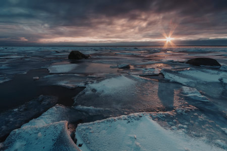 Frozen lake with melting ice floes and rocks visible, sunrise in the background, creating a serene winter landscape with clouds and water reflecting the morning light.