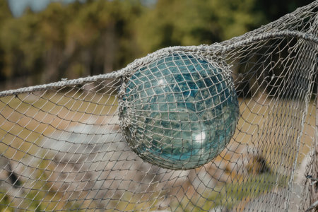 Vintage aqua glass fishing float encased in protective netting hangs outdoors near lush greenery suggesting maritime history or decoration.