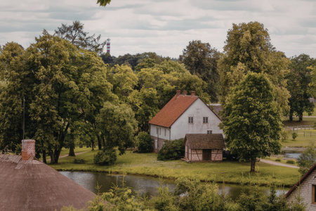 White house with red roof and small pond surrounded by lush green trees and grass in a serene countryside landscape.