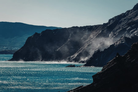 Dramatic dark rock formations on a wild coastline are continuously pounded by strong turquoise ocean waves, generating captivating white sea foam and mist.