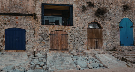 Old stone building features blue and wooden doors, balcony on rustic exterior wall with rough texture and weathered appearance, symbolizing age and character.