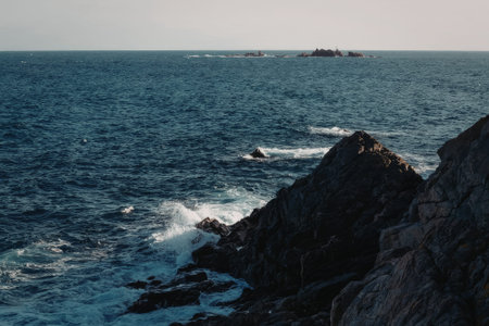 a rocky shoreline with waves crashing against the rocks, creating white foam on a sunny day with a clear blue sea and a distant island