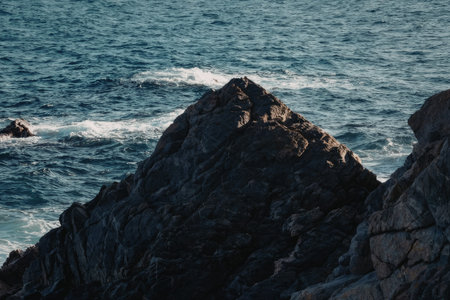 a large rock formation in the ocean with waves crashing against it. the rock is dark gray and has a rough texture. the water is blue and choppy. there are small waves crashing against the rock.