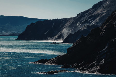 Waves crash against rugged cliffs and rocky shoreline on a clear sunny day with distant mountains visible in the serene ocean landscape.