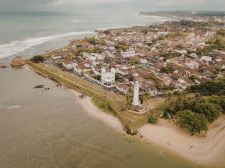 a small coastal town with a lighthouse and many houses is seen from above. the town is surrounded by greenery and has a sandy beach. the sky is overcast.