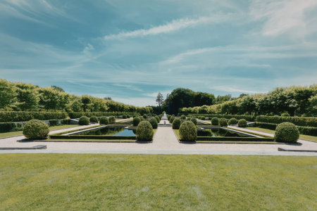 a large green field with a pathway and trees in the background. the sky is blue with some clouds. there are several bushes and trees in the image. the pathway is made of stone and has a fountain in the center.