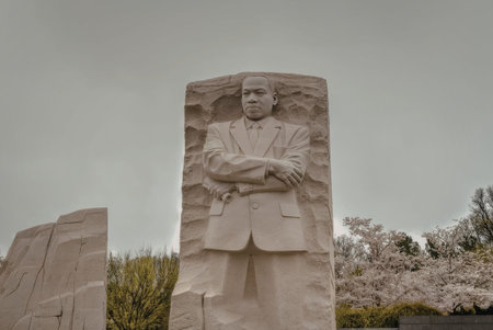 a large stone statue of a man with his arms crossed standing in front of a cloudy sky. the statue appears to be made of gray stone and has a rough texture. the man's facial expression is not visible due to the pixelation of the image. there are some green plants in the background.