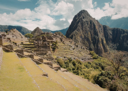 A scenic view of Machu Picchu with ancient stone ruins and terraces, surrounded by lush greenery and majestic mountains under a cloudy sky.