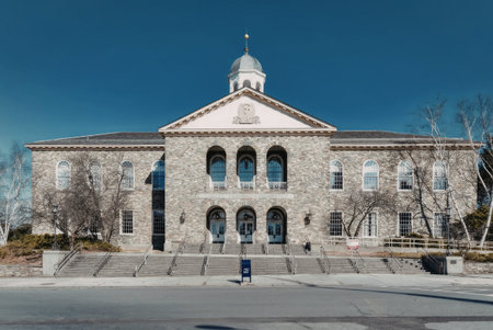 a large gray stone building with a triangular roof and a clock tower on top, the building has several arched windows and doors, and a staircase leading up to the entrance, there are trees on either side of the building and a blue sky in the background