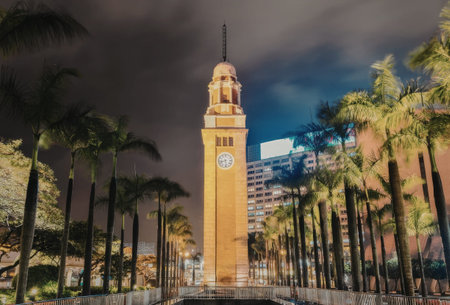 a tall yellow clock tower with a clock on the face of it stands in a park with trees and a fence