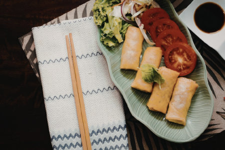 A plate of spring rolls served with sliced tomatoes and herbs, accompanied by chopsticks on a decorative napkin.