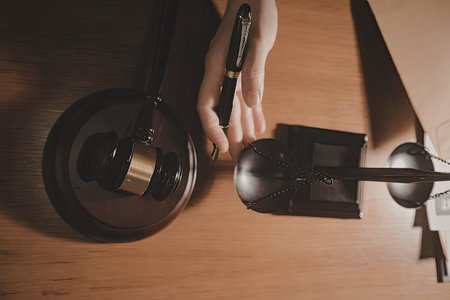 a person's hand holding a black pen over a black gavel with a gold band on a dark wooden sound block and a black sound block with a chain on a wooden desk