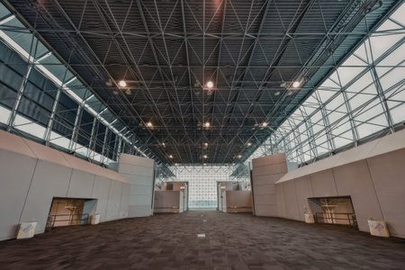 a large empty room with a high ceiling and metal beams. the walls are light gray and the floor is dark gray. there are several openings in the walls, possibly doors or windows. the ceiling is made up of metal beams and has several lights shining down. the overall atmosphere is one of emptiness and industrial functionality.