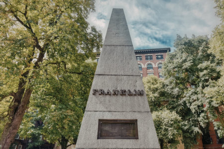 a tall gray monument with a plaque on the front is surrounded by trees and a building in the background