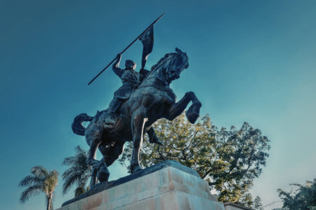 a statue of a man on horseback holding a spear in the air, with trees and a blue sky in the background