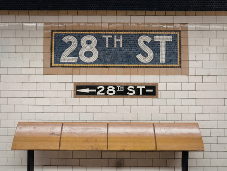 a subway station sign and bench in a tiled wall with a blue and white mosaic sign reading 28th st and a wooden bench