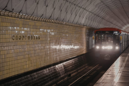 a subway train is pulling into a station with a tiled wall and a curved roof. the train has its headlights on and is on the right side of the image. the wall is made of shiny yellow tiles and has a black base. there is a sign on the wall that says cÐ¿Ð¾Ñ€Ñ‚Ð¸Ð²Ð½Ð°Ñ in gold letters.