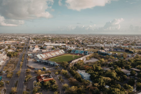 an aerial view of a city with a large stadium in the center. the stadium has a green field and colorful stands. there are several buildings and trees surrounding the stadium, and a road runs along the left side of the image.