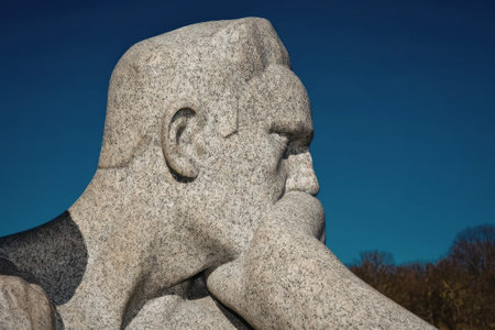 a large stone statue of a man with his hand on his chin, looking to the right. the statue is made of gray granite and has a detailed texture. the background is a clear blue sky with some trees in the distance.
