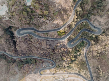an aerial view of a winding road that traverses through a mountainous area with trees and greenery on both sides of the road. the road has several sharp turns and curves, and there are a few buildings and a swimming pool visible in the top left corner of the image.