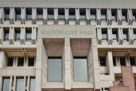 A close-up view of the Boston City Hall building, showcasing its unique modern architecture and design. The building features a predominantly concrete structure with numerous windows and a distinctive red brick section at the bottom right corner. The prominent inscription 'BOSTON CITY HALL' is carved into the stone above the entrance.