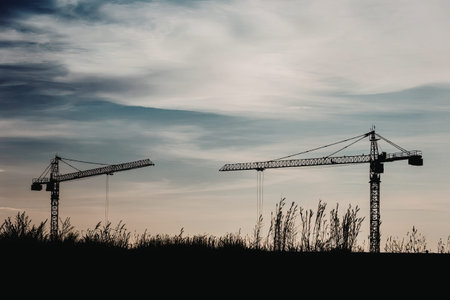 Two construction cranes stand against a serene evening sky with wispy clouds, silhouetted against the fading light.