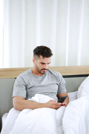 Handsone young Caucasian man spend lifestyle with smartphone and online in sicial media on website on the bed in bedroom on holiday at home. Technology and social distancing Conceptの写真素材