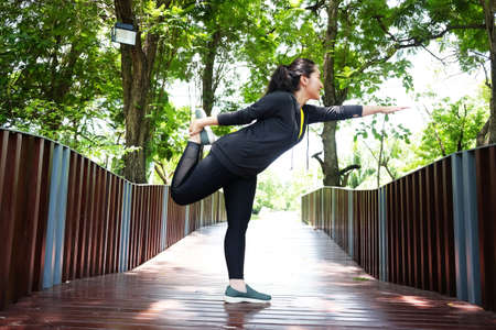 Smiling young Asian woman standing on wooden bridge for exercising and stretching the legs with Yoga...