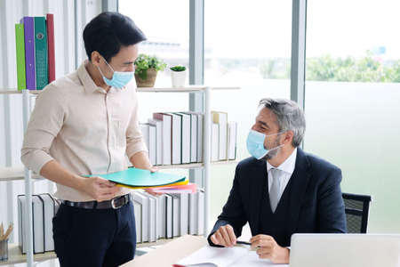 Senior Caucasian boss and young Asain businessman wearing mask preventing for Covid 19 virus holding folder work sheet and talking about job in office. Social distance and new normal lifestyle conceptの写真素材