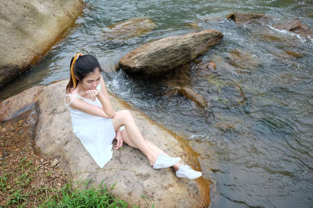 Young Asian girl sitting wearing white dress alone on the rock beside the stream in tropical rainforest.の写真素材